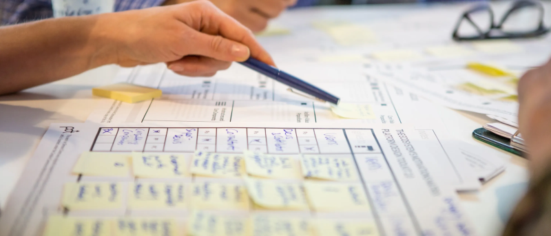 Zoomed in view of someone preparing a report. Someone is reaching across the papers with a pen.