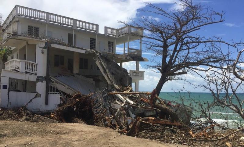 A three story white house has been partially knocked over by a large tree.